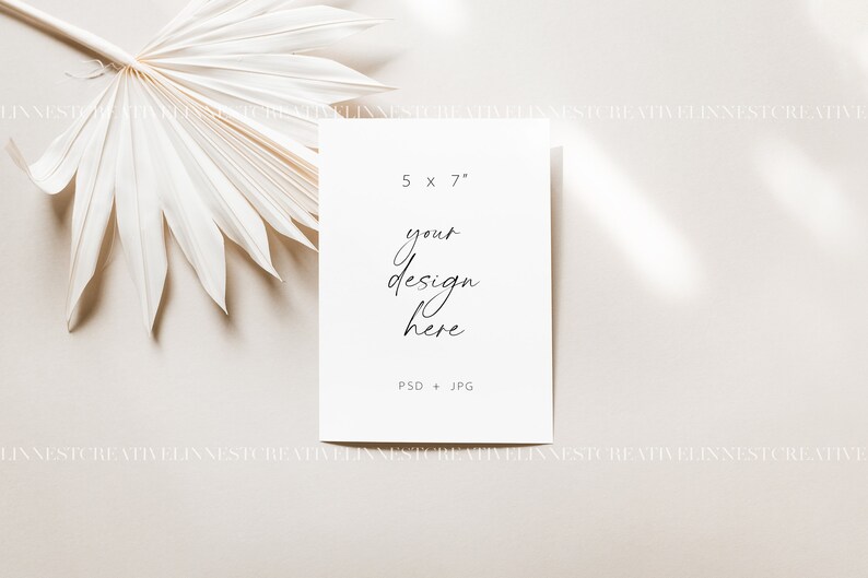 Pu&ograve; includere: Un mockup di carta bianca con il testo "5 x 7" e "your design here" su uno sfondo beige con una foglia di palma secca nell'angolo in alto a sinistra.
