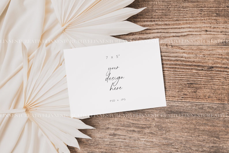 May include: A white card mockup with the text "7 x 5" and "your design here" on a wooden background with two white palm leaf fans.