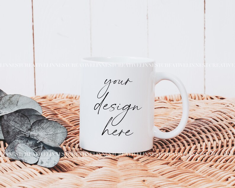 K&ouml;nnte beinhalten: Wei&szlig;e Keramiktasse mit einer schwarzen Aufschrift "your design here" auf einer geflochtenen Weidenoberfl&auml;che mit gr&uuml;nen Bl&auml;ttern.