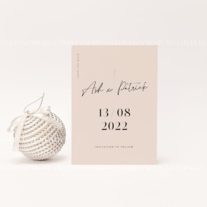以下が含まれることがあります： 薄いピンク色の「Save the Date」カードで、黒い文字が書かれています。テキストには、「Save the Date」、「Ash x Patrick」、「13/08 2022」、「Invitation to follow」とあります。