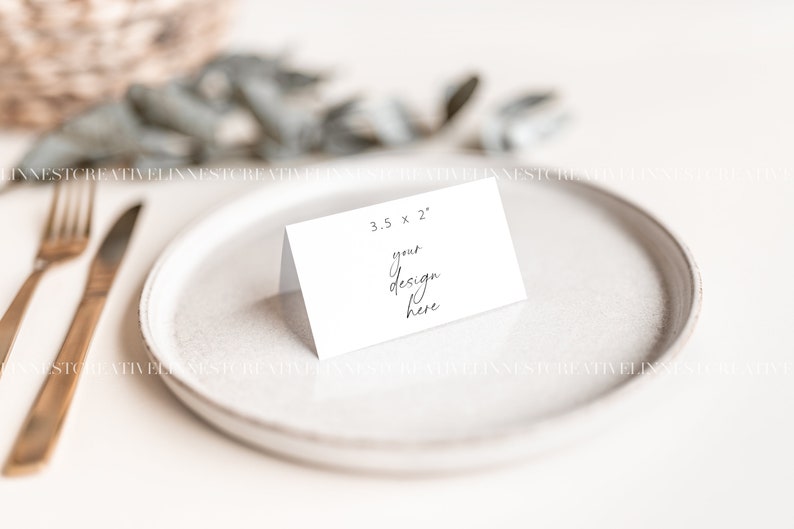 May include: A white place card with the text "3.5 x 2" and "your design here" on a white plate with gold silverware.
