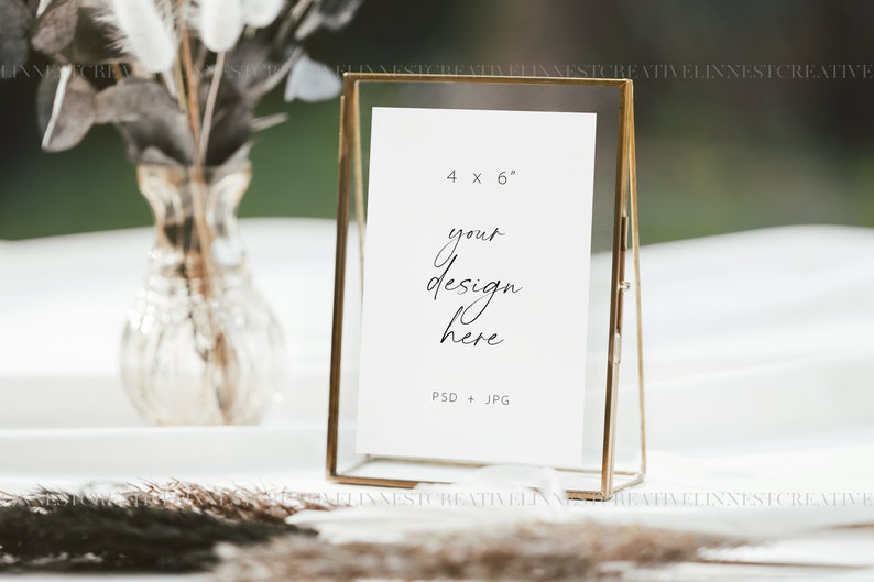 Puede incluir: Un marco de fotos de vidrio dorado con una tarjeta blanca en su interior. La tarjeta tiene el texto "4 x 6" y "tu dise&ntilde;o aqu&iacute;" escrito en ella. El marco est&aacute; sentado sobre un mantel blanco con un jarr&oacute;n de flores secas en el fondo.