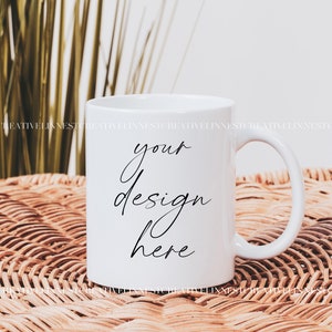 K&ouml;nnte beinhalten: Wei&szlig;e Keramiktasse mit einer schwarzen Aufschrift "your design here" auf einer geflochtenen Weidenoberfl&auml;che.