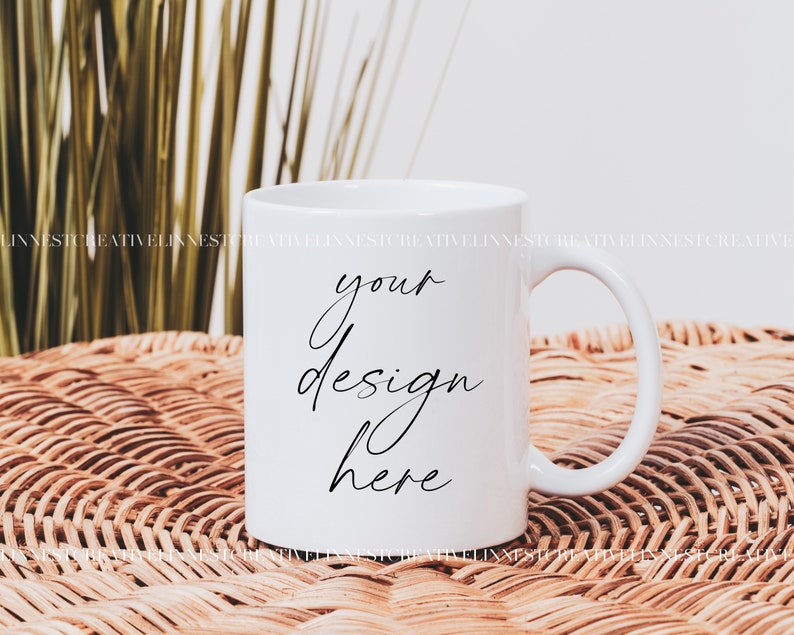 K&ouml;nnte beinhalten: Wei&szlig;e Keramiktasse mit einer schwarzen Aufschrift "your design here" auf einer geflochtenen Weidenoberfl&auml;che.