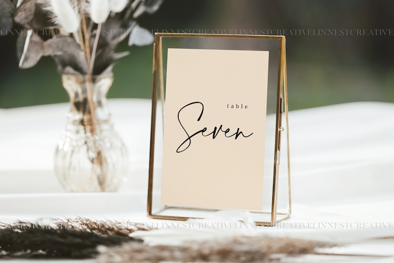 Puede incluir: Un marco dorado con una tarjeta beige en su interior. La tarjeta tiene texto negro que dice "table Seven".