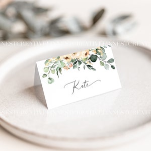 May include: A white place card with a floral design featuring yellow and peach flowers and green leaves. The name "Kate" is written in cursive on the card.