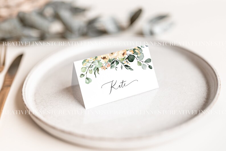 May include: A white place card with a floral design featuring yellow and peach flowers and green leaves. The name "Kate" is written in cursive on the card.
