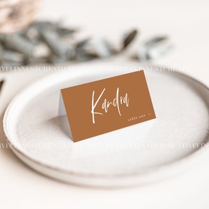 May include: A brown place card with white text that reads "Kandra table one". The place card is sitting on a white plate.