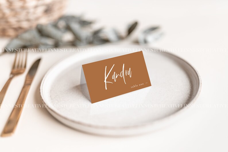 May include: A brown place card with white text that reads "Kandra table one". The place card is sitting on a white plate.