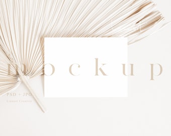 May include: A white card mockup with the word "mockup" in a light brown font, placed on a beige background with a dried palm leaf fan in the top left corner.