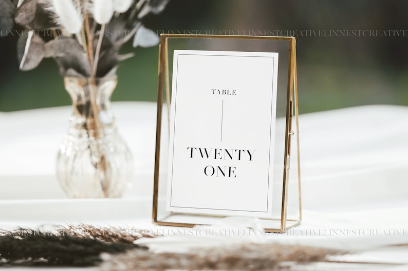 Puede incluir: Un cartel de n&uacute;mero de mesa con marco dorado y una tarjeta blanca que dice "TABLE TWENTY ONE".
