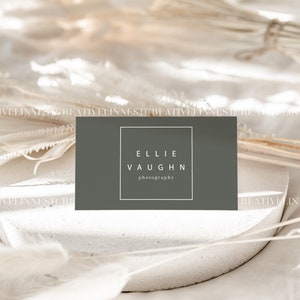 May include: A gray business card with white text that reads "Ellie Vaughn photography" on a white surface with dried white flowers.