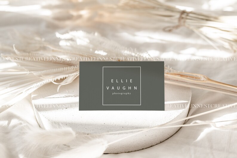 May include: A gray business card with white text that reads "Ellie Vaughn photography" on a white surface with dried white flowers.