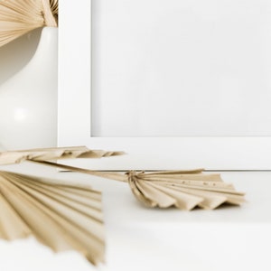 May include: A white picture frame with a blank white background.  Dried palm leaves are arranged in front of the frame on a white surface.