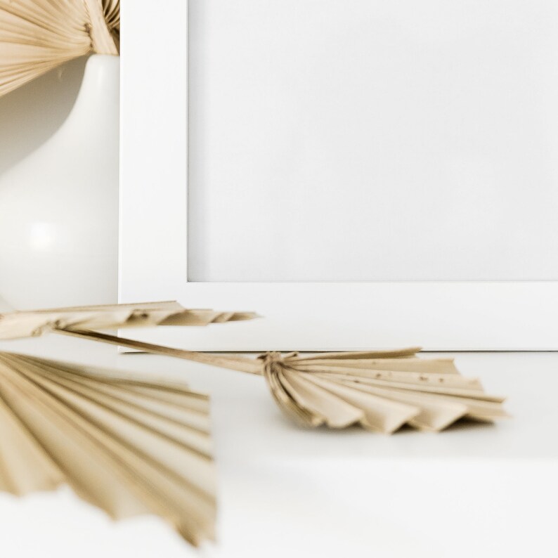 May include: A white picture frame with a blank white background.  Dried palm leaves are arranged in front of the frame on a white surface.