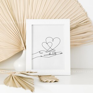 May include: A white frame with a black and white line drawing of a hand holding two intertwined hearts.
