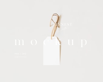 May include: A white blank gift tag with a brown string attached. The tag is hanging against a light beige background. The text "MOCKUP" is printed in white on the background.