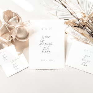 May include: Four white card mockups with text overlays, including "5 x 7", "your design here", "PSD + JPG", "5 x 3.5", and "your design here". The cards are arranged on a white surface with a pair of beige fabric baby booties and a dried floral arrangement.