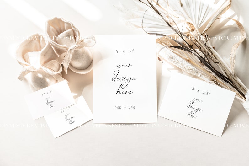 May include: Four white card mockups with text overlays, including "5 x 7", "your design here", "PSD + JPG", "5 x 3.5", and "your design here". The cards are arranged on a white surface with a pair of beige fabric baby booties and a dried floral arrangement.