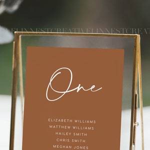 Puede incluir: Una tarjeta de n&uacute;mero de mesa marr&oacute;n con la palabra "One" escrita en cursiva blanca. La tarjeta enumera los nombres de los invitados asignados a la mesa uno: Elizabeth Williams, Matthew Williams, Hailey Smith, Chris Smith y Meghan Jones.