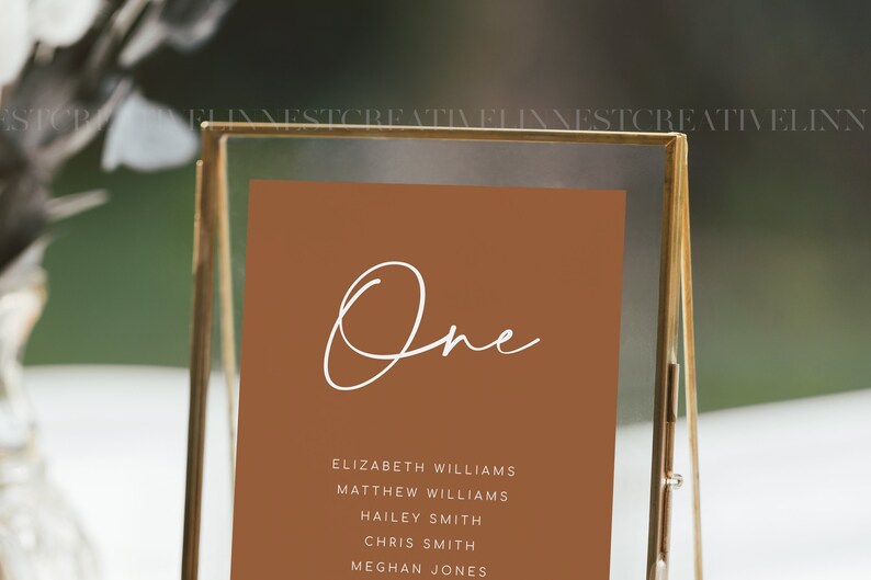 Puede incluir: Una tarjeta de n&uacute;mero de mesa marr&oacute;n con la palabra "One" escrita en cursiva blanca. La tarjeta enumera los nombres de los invitados asignados a la mesa uno: Elizabeth Williams, Matthew Williams, Hailey Smith, Chris Smith y Meghan Jones.