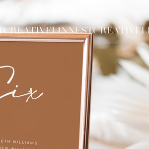 May include: A rose gold framed table number sign with the word "Six" written in white cursive script. The sign is on a white surface.