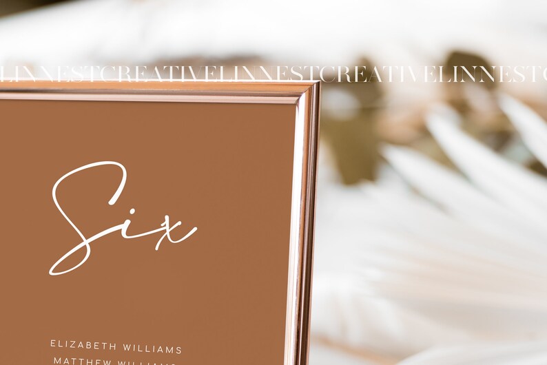 May include: A rose gold framed table number sign with the word "Six" written in white cursive script. The sign is on a white surface.