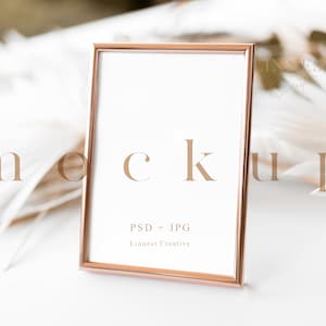 May include: A rose gold picture frame mockup with a white background. The frame is sitting on a white surface with white feathers in the background. The text "PSD + JPG" and "Linnest Creative" is printed on the frame.