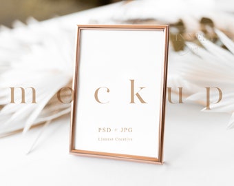 May include: A rose gold picture frame mockup with a white background. The frame is sitting on a white surface with white feathers in the background. The text "PSD + JPG" and "Linnest Creative" is printed on the frame.