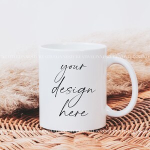 K&ouml;nnte beinhalten: Wei&szlig;e Keramiktasse mit einer schwarzen Aufschrift "your design here" auf einer geflochtenen Weidenoberfl&auml;che mit einem flauschigen wei&szlig;en Pampasgras.