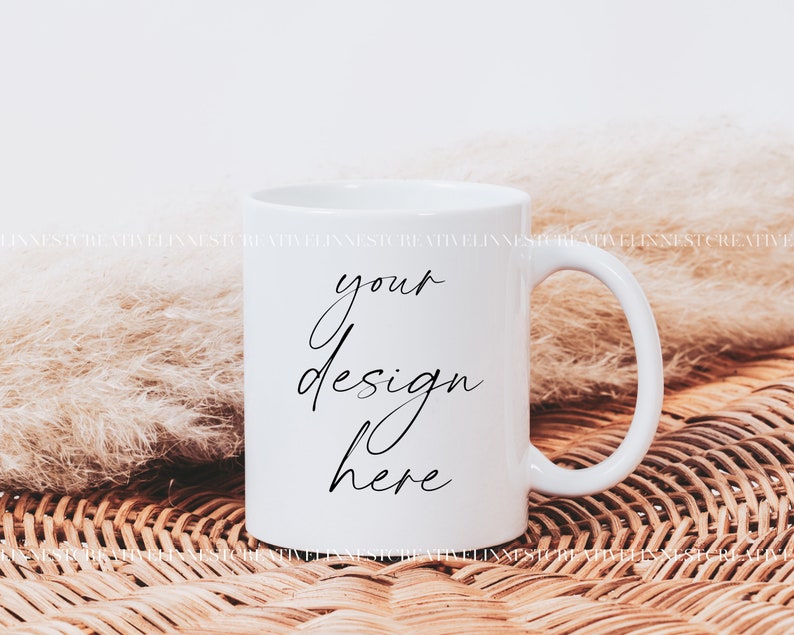 K&ouml;nnte beinhalten: Wei&szlig;e Keramiktasse mit einer schwarzen Aufschrift "your design here" auf einer geflochtenen Weidenoberfl&auml;che mit einem flauschigen wei&szlig;en Pampasgras.