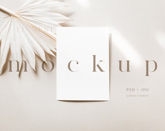 Puede incluir: Una maqueta de tarjeta blanca con la palabra "mockup" impresa en texto marrón sobre un fondo beige. Una hoja de palma seca se encuentra en la esquina superior izquierda de la imagen.
