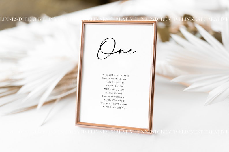 May include: A gold-framed table number sign with the word "One" written in cursive script. The sign lists the names of guests assigned to table one: Elizabeth Williams, Matthew Williams, Hailey Smith, Chris Smith, Meghan Jones, Sally Evans, Eva Montgomery, Harry Edwards, Sandra Stevenson, and Kevin Stevenson.