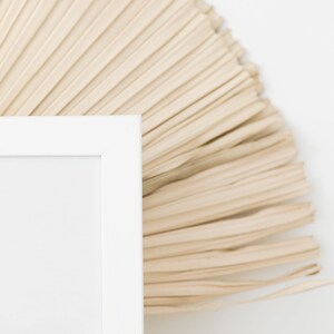 May include: A white picture frame with a blank white mat, sitting in front of a beige woven fan.