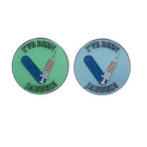 May include: Two round pins with a green and blue background. The pins feature a white bandage and a blue syringe with the text "I've Been Jabbed!" in black.