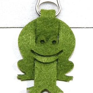 Peut inclure: Tirette de fermeture éclair en forme de grenouille verte en feutre, avec un visage souriant et de petits pieds. La grenouille est attachée à une fermeture éclair en métal argenté. Accessoire amusant.