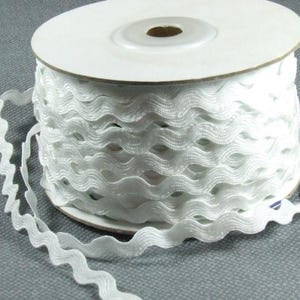 Könnte beinhalten: Eine Rolle weißes, wellenförmiges Band. Das Band besteht aus einem weichen, strukturierten Material und ist um eine Papprolle gewickelt. Die Kanten des Bandes haben ein gewelltes Design, das eine dekorative Verzierung bildet. Das Band ist ideal für Bastel- und Nähprojekte.