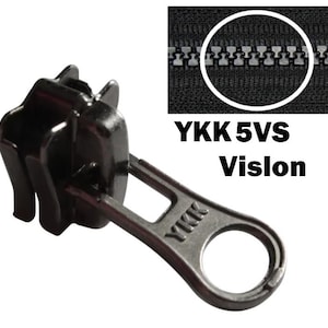 Peut inclure: Gros plan sur une tirette de fermeture éclair noire YKK 5VS Vislon. La tirette a une forme arrondie avec une boucle pour l'attache. L'image montre également un gros plan des dents de la fermeture éclair.