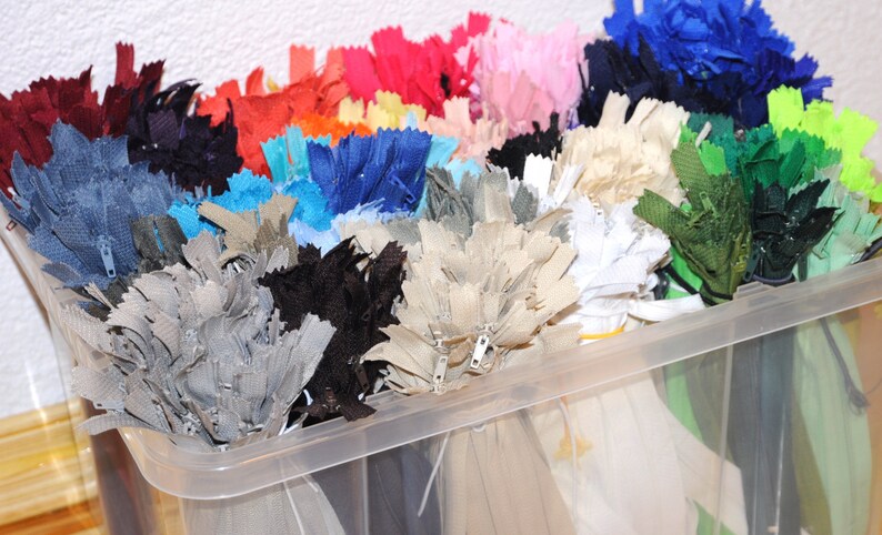 May include: A clear plastic storage container filled with a variety of colours of zips. The zips are organised by colour and are ready to be used for sewing projects.