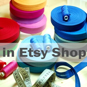 May include: Multiple rolls of colorful bias tape in red, orange, pink, light blue, and dark blue, accompanied by matching sewing thread spools and a white measuring tape with black markings showing numbers up to 150 centimeters for scale.