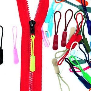 Op de afbeelding: Een rode ritssluiting met een zwarte ritstrekker en een verzameling kleurrijke ritstrekkers. De ritstrekkers zijn in verschillende kleuren, waaronder blauw, zwart, roze, groen en bruin.