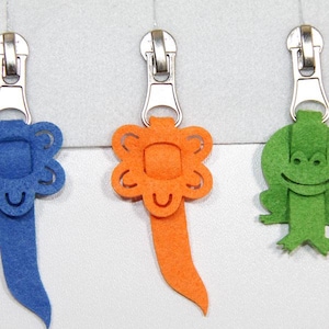 Könnte beinhalten: Drei Reißverschlussanhänger aus Filz mit verschiedenen Designs. Der erste ist eine blaue Blume mit einem lächelnden Gesicht, der zweite ist eine orangefarbene Blume mit einem lächelnden Gesicht und der dritte ist ein grüner Frosch mit einem lächelnden Gesicht.