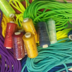 May include: A collection of colorful sewing supplies including multiple spools of thread in red, yellow, dark blue, light blue, and pink, alongside coiled lengths of purple, lime green, yellow, and teal cord, with a measuring tape visible.