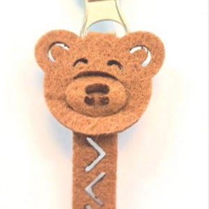 Peut inclure: Tirette de fermeture éclair en feutre marron avec un motif de visage d'ours. L'ours a un nez et une bouche cousus, avec une étiquette rectangulaire en dessous. L'étiquette a un motif de couture en zigzag. La tirette est attachée à une fermeture éclair en métal argenté.