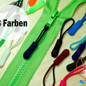 Op de afbeelding: Een close-up van een groene rits met diverse gekleurde ritstrekkers. De afbeelding toont een verscheidenheid aan kleuren, waaronder blauw, zwart, rood en groen. De tekst "18 Farben" staat in een witte cirkel.