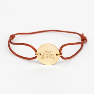 May include: A brown cord bracelet with a gold circular charm that says "Not Today Satan" in a retro font.