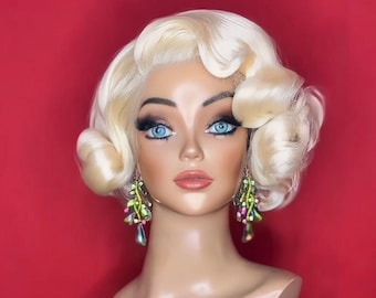 May include: A blonde wig with short, curled hair styled in a vintage bob. The wig is on a mannequin head with a light brown skin tone. The mannequin is wearing green and pink dangle earrings.