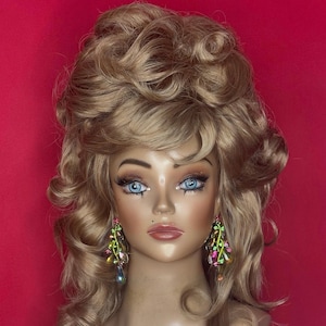 May include: A mannequin head with a blonde wig styled in voluminous curls and a bouffant. The mannequin has blue eyes, full makeup, and is wearing colorful, dangling earrings. The background is a solid red.