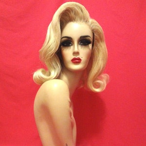 Peut inclure: Une perruque blonde avec un style volumineux et bouclé. La perruque est coiffée avec un bouffant haut et présente des boucles douces et fluides. La perruque est présentée sur une tête de mannequin sur un fond rouge.
