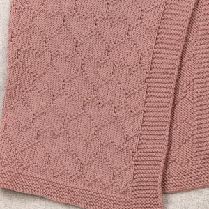 May include: A dusty rose knitted blanket featuring a repeating heart pattern throughout the main body and a wide, textured garter stitch border along the edges. The fabric has a consistent, soft-looking knit construction with a uniform color.